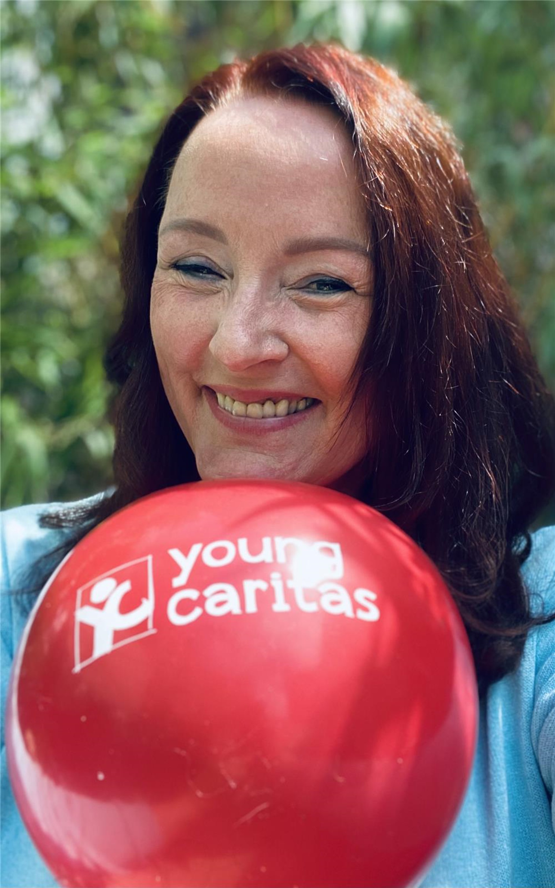 Frau mit rotbraunen Haaren hält youngcaritas Ballon und lächelt in die Kamera